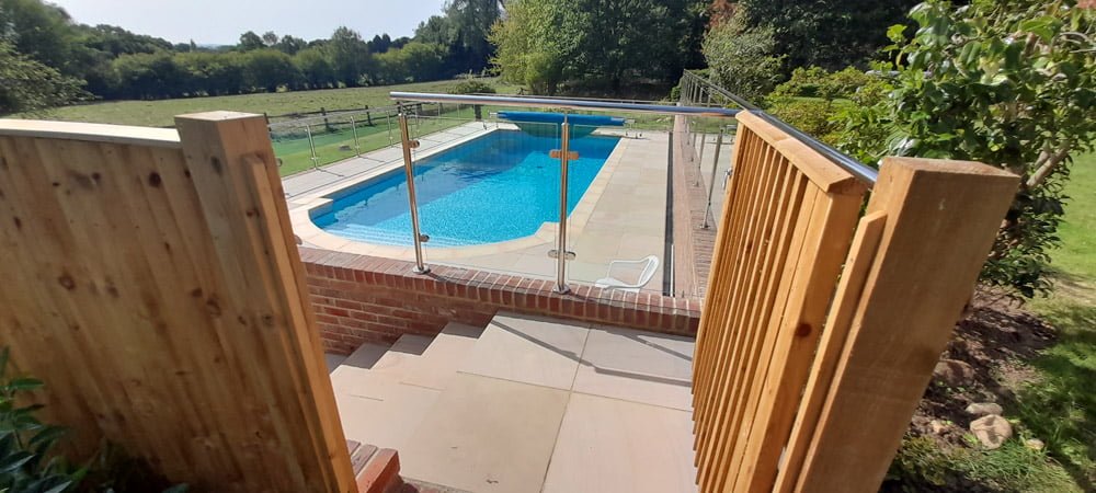 Outdoor swimming pool surrounded by patio and fields. There are also steps down to the pool with a glass fenced off area