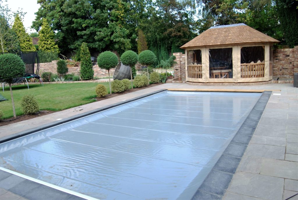 Grey swimming pool cover on an outdoor pool with a wooden outbuilding and plants surrounding