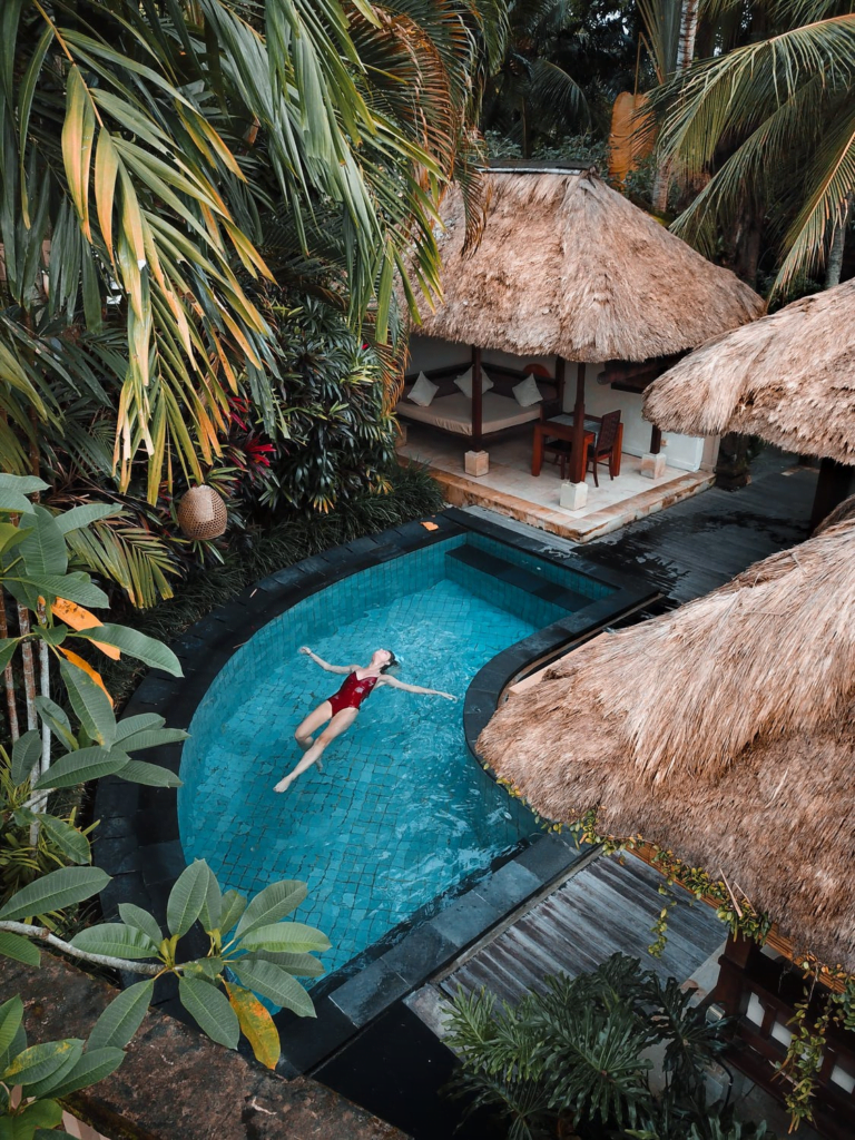 Woman floating in a shaped outdoor swimming pool surrounded by outbuilding with sunbeds