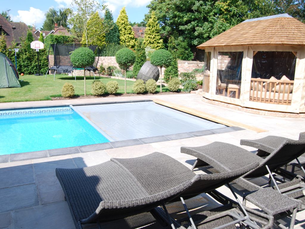A grey swimming pool cover half covering a swimming pool surrounded by sunbeds and an outbuilding